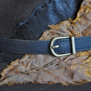 May include: A brown leather hat with a black leather band and a brass buckle. The hat is decorated with a piece of brown leather that has been stitched on.