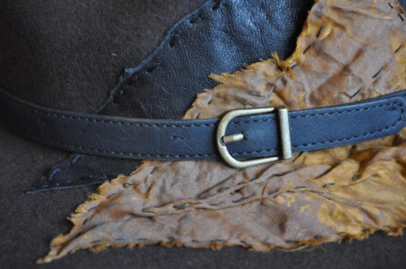 May include: A brown leather hat with a black leather band and a brass buckle. The hat is decorated with a piece of brown leather that has been stitched on.