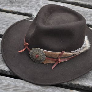 May include: A brown felt cowboy hat with a brown leather band and a metal concho. The hat has a white feather trim.