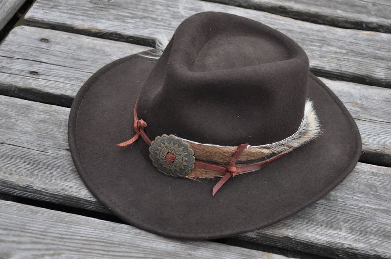 May include: A brown felt cowboy hat with a brown leather band and a metal concho. The hat has a white feather trim.