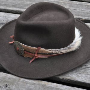 May include: A brown felt fedora hat with a wide brim and a decorative band made of leather, feathers, and a metal flower accent.