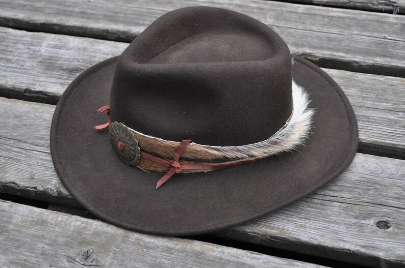 May include: A brown felt fedora hat with a wide brim and a decorative band made of leather, feathers, and a metal flower accent.