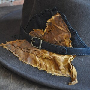 May include: A brown felt fedora hat with a black leather band and a brown leather patch sewn on the front. The patch has a frayed edge and is stitched with brown thread.