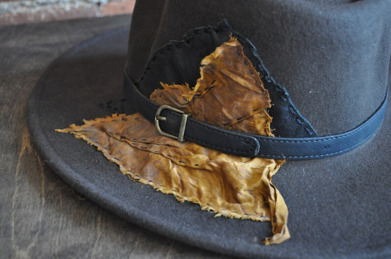 May include: A brown felt fedora hat with a black leather band and a brown leather patch sewn on the front. The patch has a frayed edge and is stitched with brown thread.