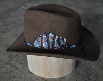 May include: A brown felt cowboy hat with a wide brim and a decorative band of blue and white triangular pieces.