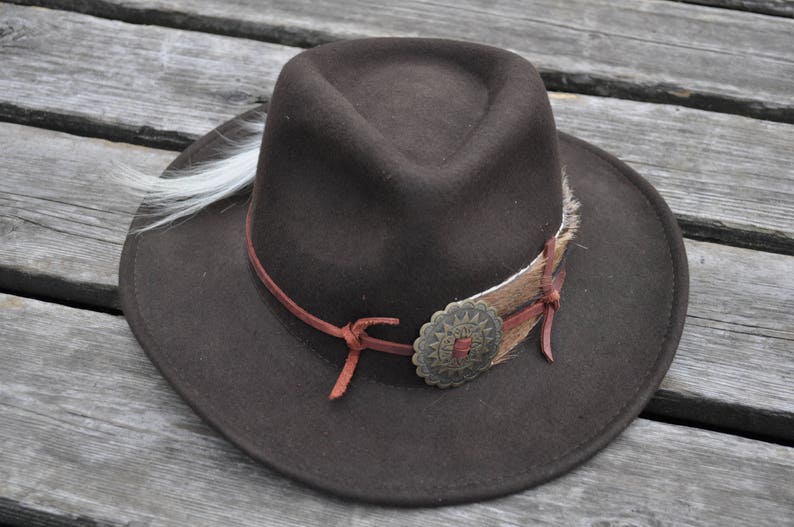 May include: A brown felt cowboy hat with a brown leather band and a metal concho. The hat has a white feather trim.