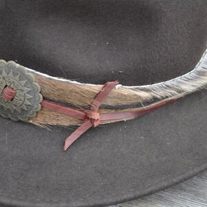 May include: A brown felt cowboy hat with a brown leather band and a fur trim. The hat has a decorative metal concho with a starburst design.