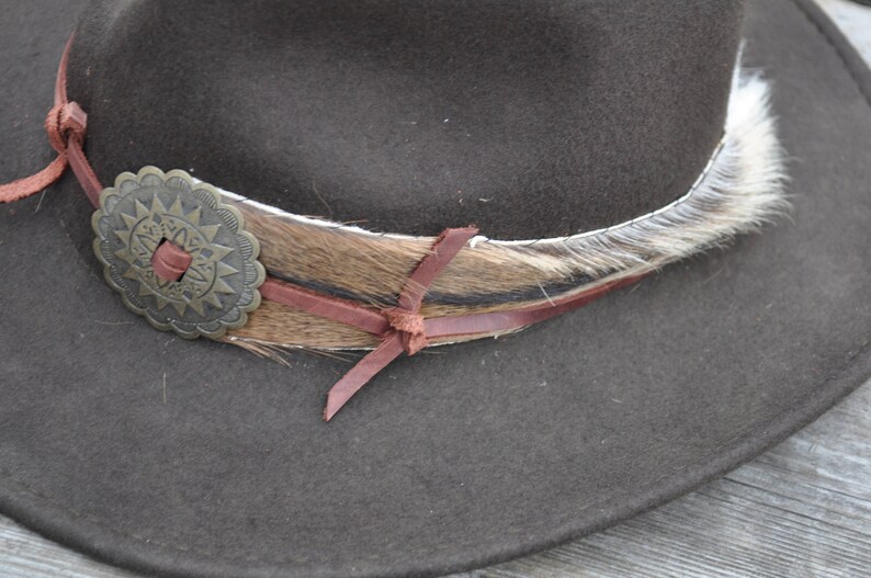 May include: A brown felt cowboy hat with a brown leather band and a fur trim. The hat has a decorative metal concho with a starburst design.