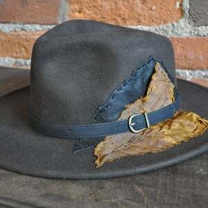 May include: A brown felt fedora hat with a wide brim and a leather band. The hat has a unique design featuring a large, brown leaf attached to the crown.