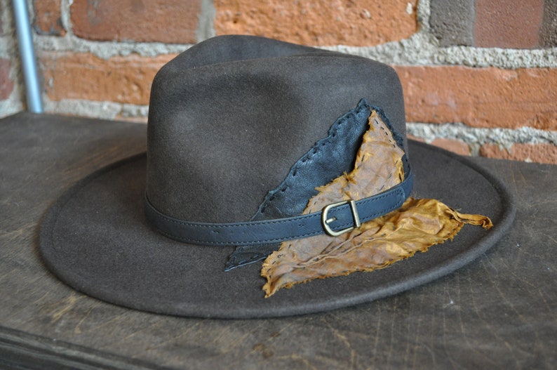 May include: A brown felt fedora hat with a wide brim and a leather band. The hat has a unique design featuring a large, brown leaf attached to the crown.