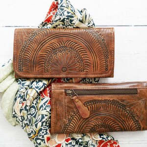 Puede incluir: Dos carteras de cuero marr&oacute;n con intrincados dise&ntilde;os florales. Las carteras est&aacute;n apiladas una encima de la otra y est&aacute;n sobre una superficie blanca con una tela estampada debajo.