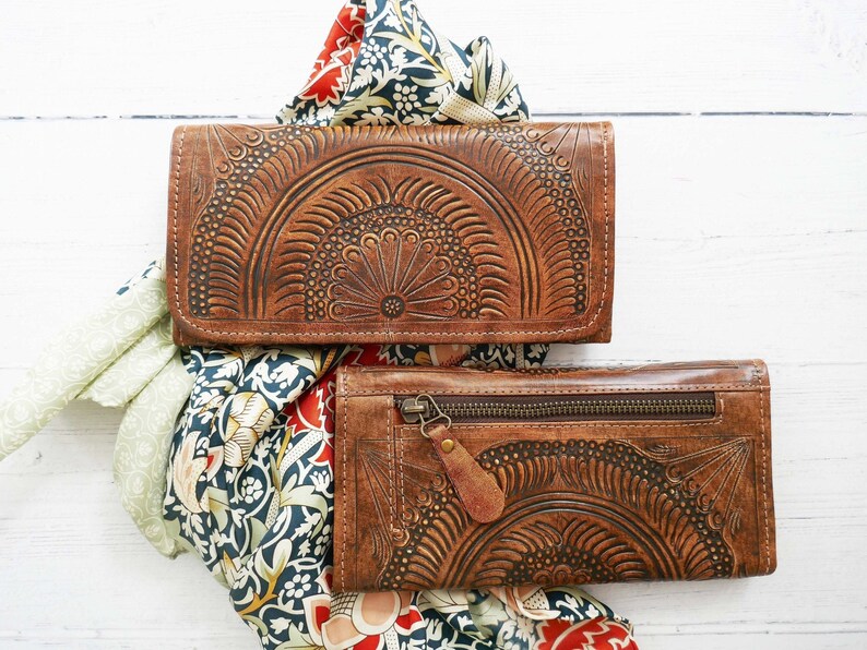 Puede incluir: Dos carteras de cuero marr&oacute;n con intrincados dise&ntilde;os florales. Las carteras est&aacute;n apiladas una encima de la otra y est&aacute;n sobre una superficie blanca con una tela estampada debajo.