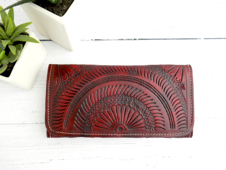 Puede incluir: Una cartera de cuero burdeos con intrincados dise&ntilde;os florales en relieve. La cartera tiene un borde cosido y un cierre de solapa.