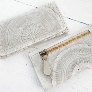 Op de afbeelding: Twee off-white leren portemonnees met reliëf bloemen en geometrische patronen. Eén portemonnee is opgevouwen met een drukknoopsluiting, de andere heeft een gouden rits. De portemonnees liggen op een wit houten oppervlak.