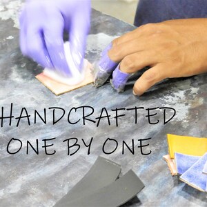 Puede incluir: Una mano con guantes morados aplica una sustancia blanca a un trozo de cuero. El cuero est&aacute; rodeado de otros trozos de cuero de varios colores, incluyendo amarillo, azul y rosa. El texto "HANDCRAFTED ONE BY ONE" est&aacute; escrito en negro sobre una superficie gris.