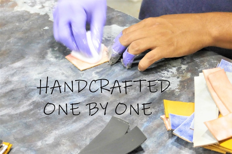 Puede incluir: Una mano con guantes morados aplica una sustancia blanca a un trozo de cuero. El cuero est&aacute; rodeado de otros trozos de cuero de varios colores, incluyendo amarillo, azul y rosa. El texto "HANDCRAFTED ONE BY ONE" est&aacute; escrito en negro sobre una superficie gris.