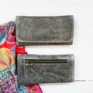 Puede incluir: Dos carteras de cuero gris con un patr&oacute;n decorativo en relieve. La cartera superior est&aacute; cerrada y la cartera inferior est&aacute; abierta, mostrando una cremallera y un tirador de cremallera dorado.