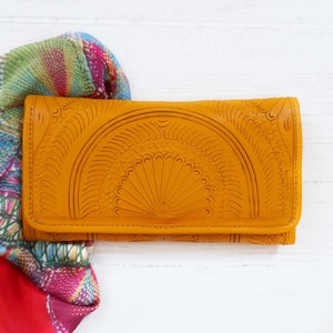 Puede incluir: Una cartera de cuero amarilla con un dise&ntilde;o en relieve intrincado. La cartera est&aacute; cerrada y se encuentra sobre una superficie blanca.