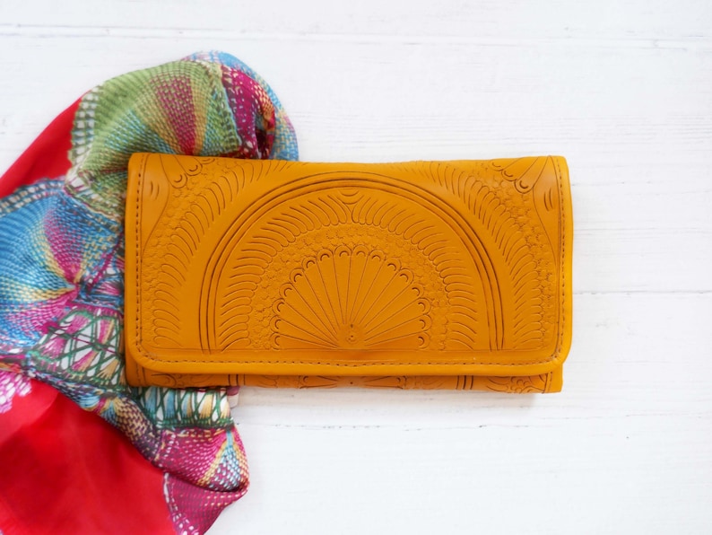 Puede incluir: Una cartera de cuero amarilla con un dise&ntilde;o en relieve intrincado. La cartera est&aacute; cerrada y se encuentra sobre una superficie blanca.