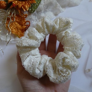May include: A white lace scrunchie, a hair tie, made of delicate white lace. The scrunchie is gathered in a circle and is held in a hand.