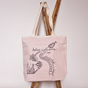 May include: A pale pink tote bag with cream handles. The bag has a black ink illustration with the words "believe in your dreams and follow your heart." The design includes a path of hearts, a floral element, and a heart-shaped arrow.