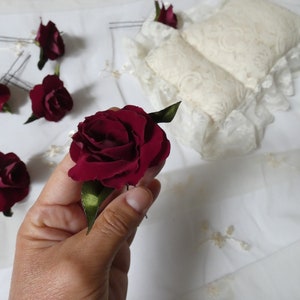 May include: Burgundy rose hair pins with green leaves. Several pins are arranged on a white surface, with a hand holding one of the roses. The background includes a lace-trimmed pillow and fabric.