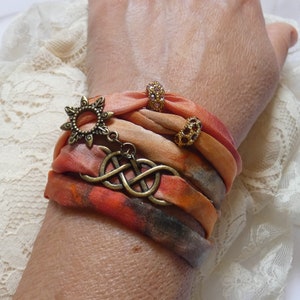 Può includere: Un braccialetto di seta multistrato con un ciondolo a forma di sole in bronzo e un pendente a nodo celtico. Il braccialetto è decorato con piccole perline scintillanti in tonalità arancio e oro.