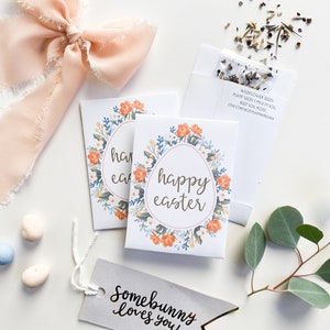 May include: A set of three small white envelopes with floral designs and the text "happy easter" inside. The envelopes are accompanied by a small white bag of wildflower seeds, a pink ribbon, and a gray tag with the text "Somebunny loves you!" in gold lettering.