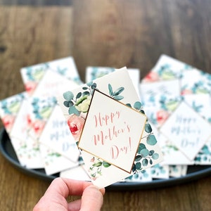 May include: A hand holding a white Mother's Day card with the text "Happy Mother's Day!" in a gold geometric frame. The card is decorated with watercolor floral and eucalyptus designs. Other cards are on a dark gray tray.