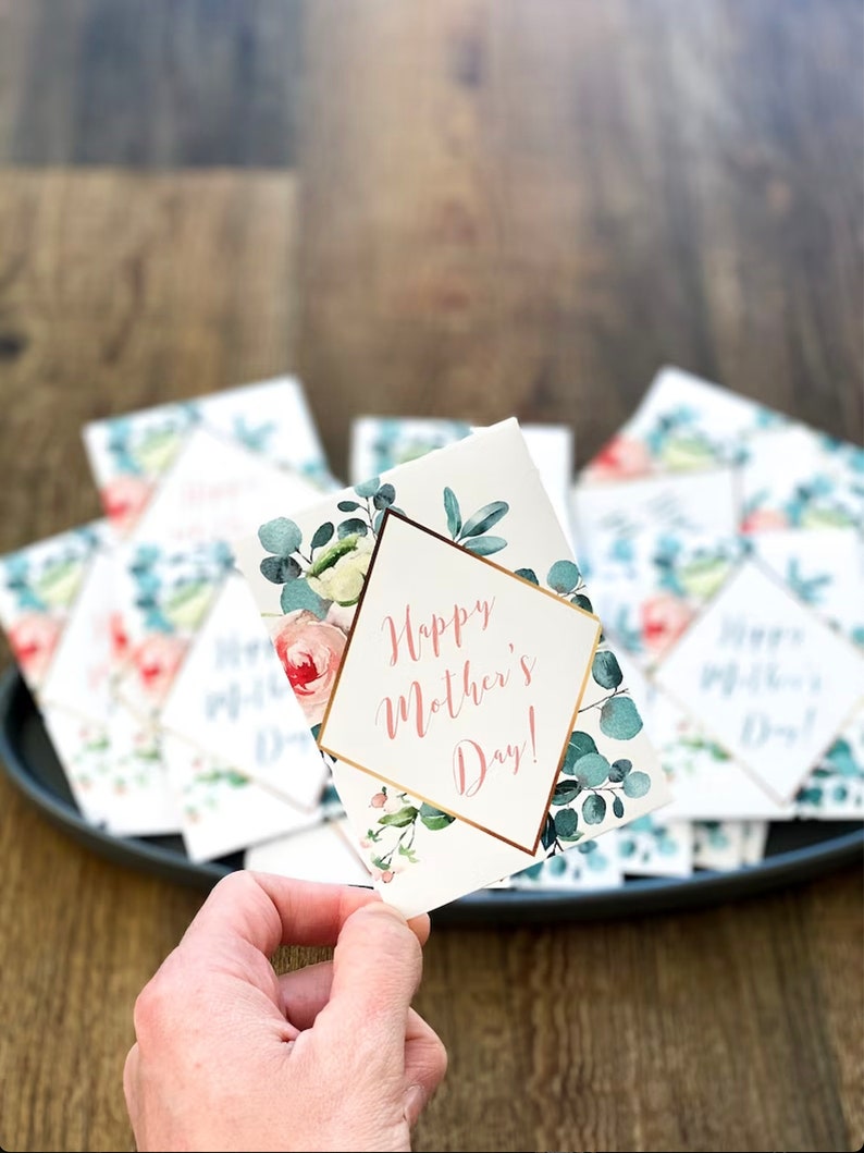 May include: A hand holding a white Mother's Day card with the text "Happy Mother's Day!" in a gold geometric frame. The card is decorated with watercolor floral and eucalyptus designs. Other cards are on a dark gray tray.