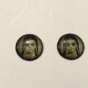 May include: Two round, black-rimmed buttons with a vintage-style image of a young girl with dark hair and a white dress. The girl has a serious expression and is looking directly at the viewer.