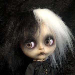 Puede incluir: Muñeca hecha a mano con tez pálida, ojos marrones grandes y peluca bicolor en blanco y negro. La muñeca viste un vestido negro con un colgante de llave. La muñeca tiene una estética gótica o alternativa.