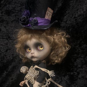 May include: A doll with curly blonde hair, wearing a black dress and holding a skeleton. The doll has large yellow eyes and a black top hat with purple accents, black roses, and a tag that reads "Funerals 10%".