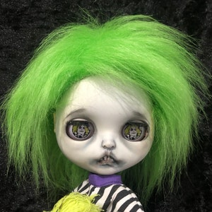 May include: A doll with green hair and a pale face. The doll has large eyes with a printed image of a character. The doll is wearing a black and white striped shirt with a purple collar and is holding a small doll with yellow hair.