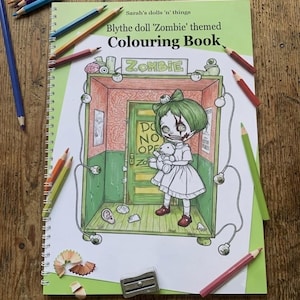 Peut inclure: Un livre de coloriage &agrave; spirales intitul&eacute; Sarah's dolls 'n' things Blythe doll 'Zombie' themed Colouring Book. La couverture pr&eacute;sente une illustration au trait d'une poup&eacute;e aux cheveux verts tenant un ours en peluche dans une pi&egrave;ce avec une porte marqu&eacute;e DO NOT OPEN.