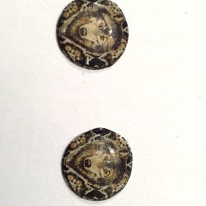 Puede incluir: Un par de pendientes redondos, negros y marrones con un diseño de cara de lobo. La cara de lobo está rodeada por un patrón geométrico.