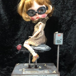 Könnte beinhalten: Eine Puppe mit blonden Haaren und einer Schutzbrille sitzt auf einem mechanischen Fliegenkarussell. Das Karussell ist grau lackiert und trägt ein Schild mit der Aufschrift "Ride The Fly" und einem Preis von 10 Cent.
