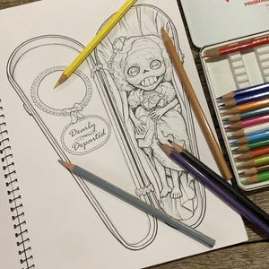 May include: A black and white line art drawing in a spiral notebook featuring a character with large eyes and teeth inside a coffin, with the text Dearly Departed. Colored pencils in yellow, tan, purple, and black are placed on the page.