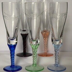 May include: A collection of six clear glass cordial glasses with etched floral patterns. Each glass features a different coloured base and stem, including blue, purple, green, orange, and light blue. The stems have a twisted rope design.