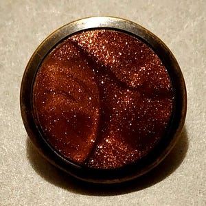 May include: A round, bronze-colored button with a shimmering, reddish-brown center. The center has a glittery, speckled appearance, with a darker, swirled pattern. The button has a raised, bronze-colored rim.