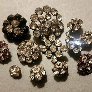 May include: A collection of vintage rhinestone brooches and earrings in various shapes and sizes. The jewelry features clear, black, and red rhinestones set in metal. The pieces include floral, round, and abstract designs, showcasing a range of styles.