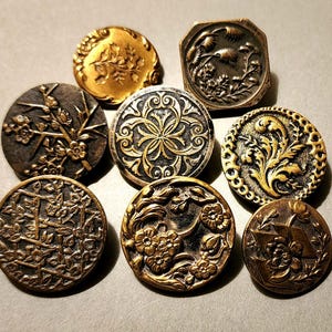 May include: A collection of antique buttons in various shapes and sizes. The buttons are made of metal and feature intricate floral and geometric designs in shades of gold, bronze, and black. Some buttons have raised relief patterns.