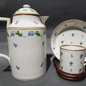 May include: A white porcelain tea set with blue floral designs and gold trim. The set includes a tall coffee pot, a cup, a saucer, and a wooden coaster. The floral pattern features blue bellflowers and green leaves.