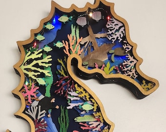 Könnte beinhalten: Eine Holzskulptur eines Seepferdchens mit einem detaillierten Ausschnittdesign. Das Seepferdchen ist mit bunten Bildern von Meereslebewesen gefüllt, darunter Fische, Korallen, ein Tintenfisch und eine Robbe. Die Skulptur ist mit LED-Lichtern beleuchtet.