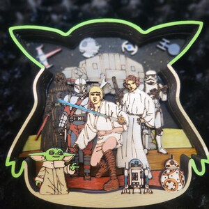 May include: A multi-layered wooden cutout featuring characters from Star Wars. The characters include Yoda, Luke Skywalker, Darth Vader, R2-D2, and a Mandalorian. The background is a dark blue space scene with stars.