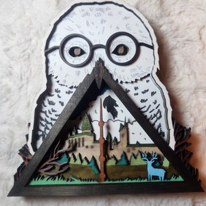 May include: A wooden owl sculpture with a black and white painted design. The owl's eyes are painted with a realistic look. The owl is holding a triangular wooden frame with a scene of a castle and a deer inside. The castle is painted in shades of brown and green, and the deer is painted in blue and white.