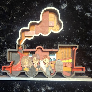 May include: A wooden 3D model of the Hogwarts Express train with Harry Potter, Ron Weasley, and Hermione Granger figures inside. The train is red and black with a brown smoke plume. The figures are detailed and painted in their signature colors.