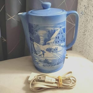 May include: A light blue ceramic electric teapot featuring a white winter landscape scene with a house, a horse-drawn carriage, and a person walking a dog. The text Homestead in Winter is printed at the base. A white power cord is included.