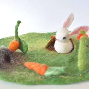 Op de afbeelding: Een wit vilt konijn kijkt uit een gat in een groen vilt veld. Twee oranje vilt wortels zijn in het veld, een met groene vilt bladeren.