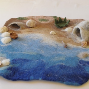 May include: A felt play mat depicting a beach scene with a blue ocean, brown sand, and a small green bush. There are several white and brown seashells scattered on the sand, along with a small brown seashell in the water.